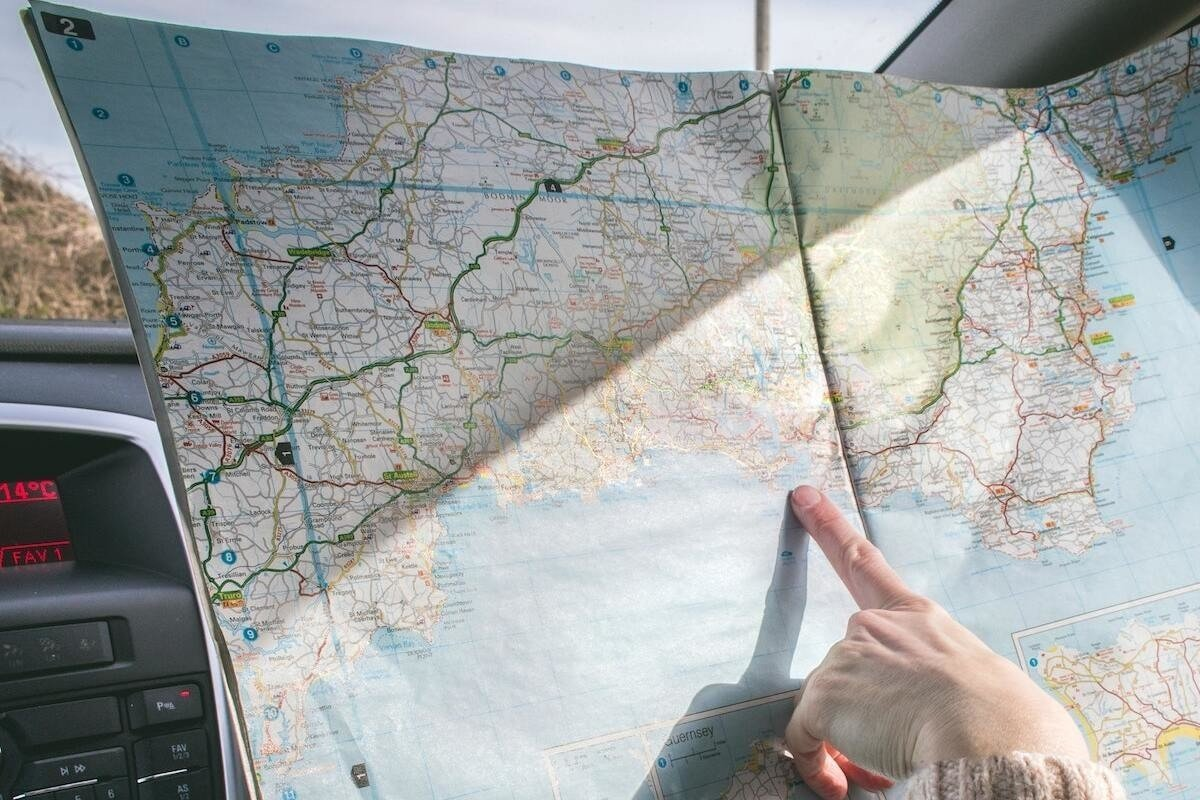 A person sits in a car, holding and pointing at a large road map spread open on the dashboard, with sunlight illuminating part of the map.