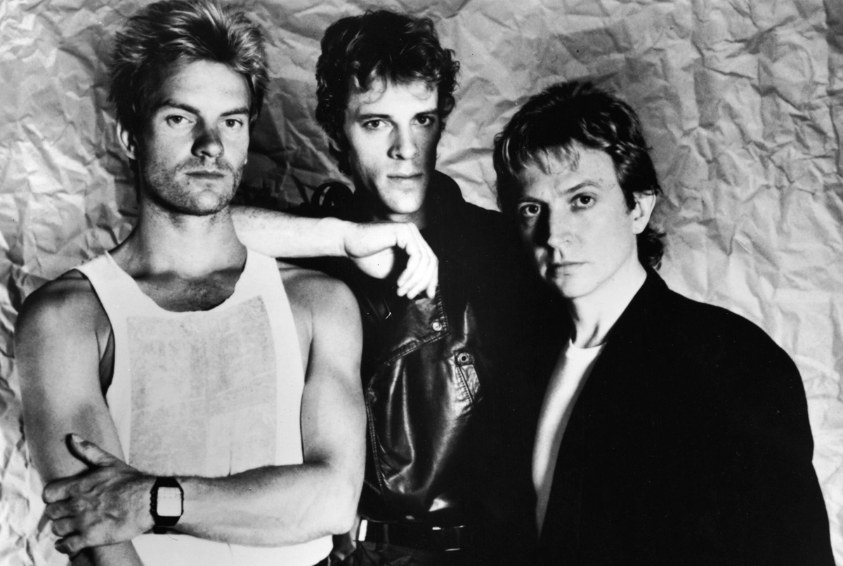 Three men pose together in front of a crumpled background. One wears a white tank top and watch, another a leather jacket, and the third a dark suit jacket. All look directly at the camera with serious expressions. song