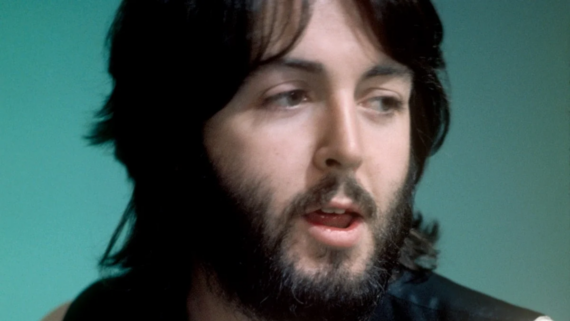 A man with medium-length dark hair and a full beard looks to the side with his mouth slightly open. He is wearing a dark vest over a light shirt, against a teal background.