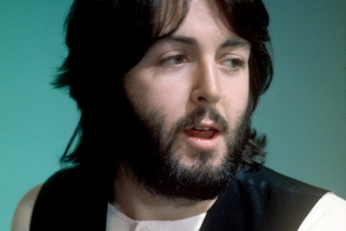 A man with medium-length dark hair and a full beard looks to the side with his mouth slightly open. He is wearing a dark vest over a light shirt, against a teal background.
