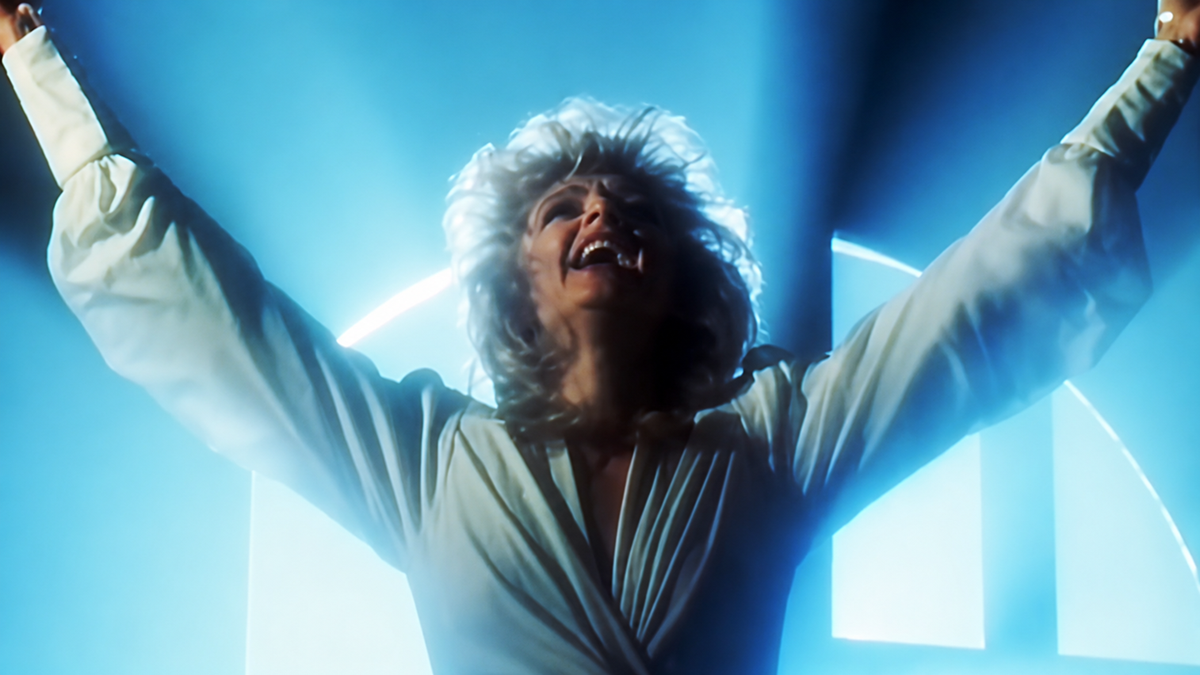 A person with light hair, wearing a white outfit, stands with arms raised in front of a glowing arched window, illuminated by blue light.