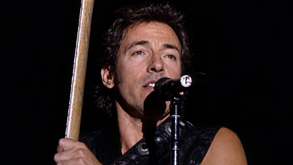 A man with wavy dark hair and a sleeveless black leather vest sings into a microphone on stage, holding a guitar neck, with a dark background.