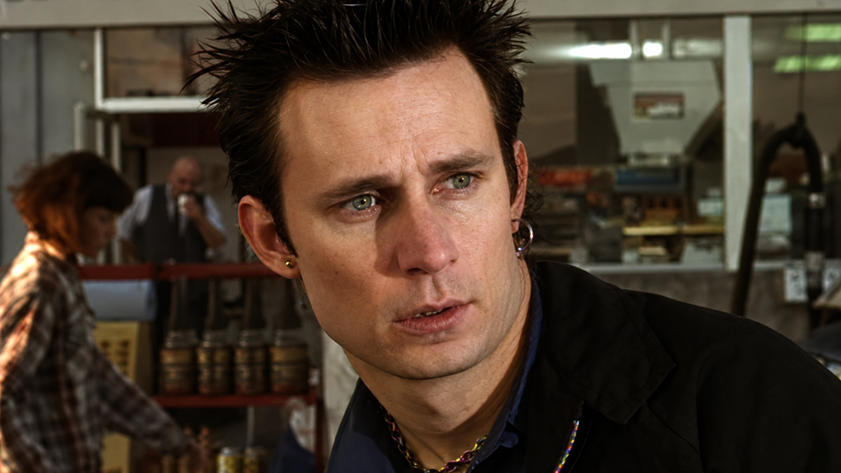 A man with spiked dark hair and earrings looks to the side with a serious expression. He is outdoors near a market or shop, with people and shelves of goods in the blurred background.