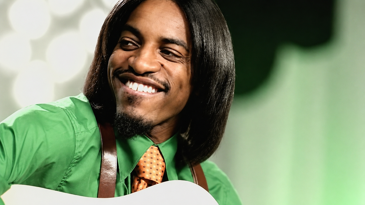 A smiling man with straight, shoulder-length hair wears a bright green shirt and orange polka-dot tie, holding a guitar. The background is softly lit with green and white tones.
