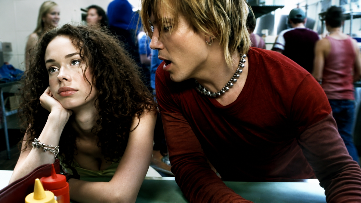 A woman with curly hair looks bored and rests her head on her hand at a diner counter, while a person with short blond hair and a spiked necklace talks to her. Other people are blurred in the background.