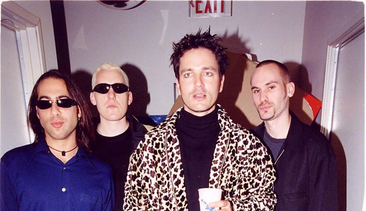 Four men stand together indoors; one in a leopard print jacket holds a cup, one wears sunglasses, and all appear at a casual gathering with streamers in the background and an exit sign overhead.