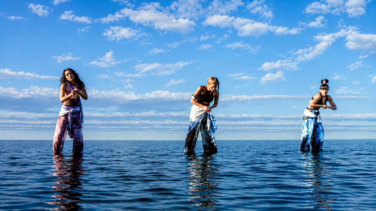 Three women in colorful clothing stand in shallow ocean water, performing expressive dance poses under a bright blue sky filled with scattered clouds.