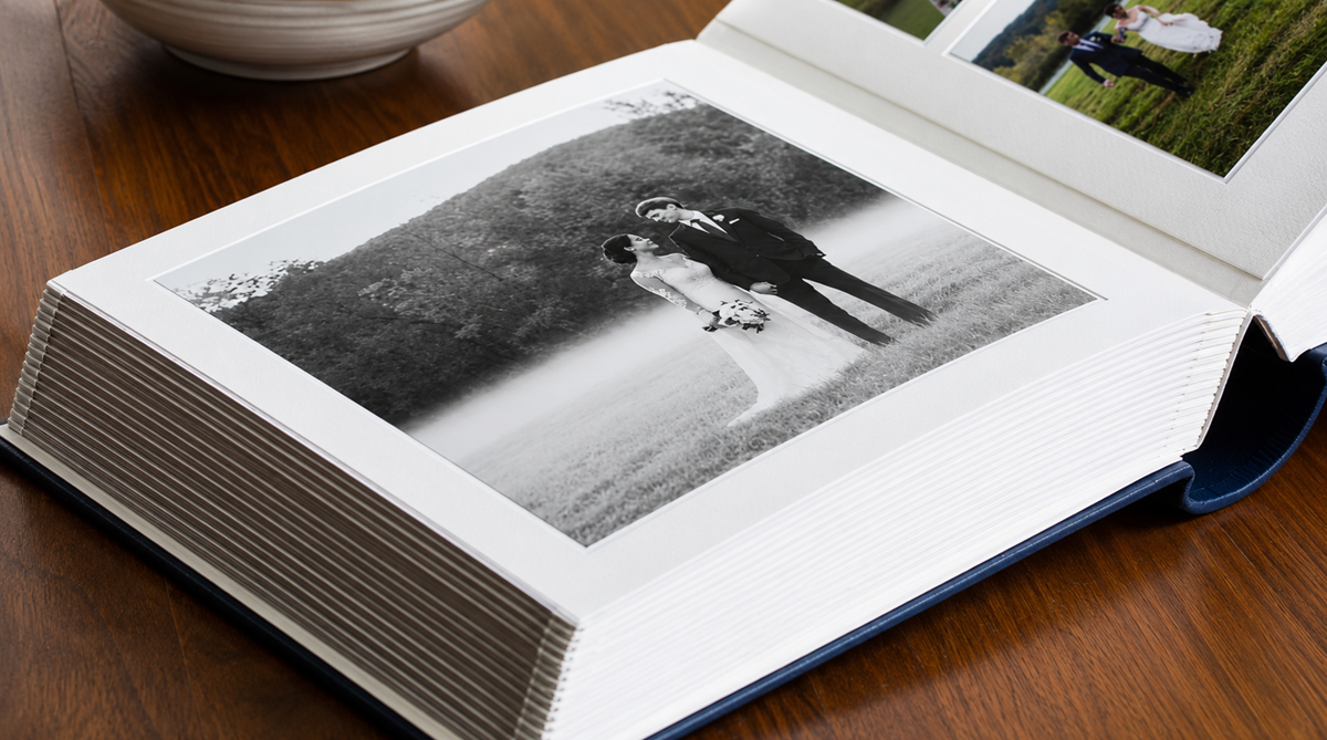 Open photo album on a wooden table, displaying a black and white wedding photo of a couple standing outdoors, the bride holding a bouquet, with trees and grass in the background.