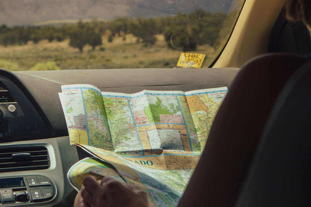A person sits in a car passenger seat holding a colorful road map, looking out at a landscape of trees and distant mountains through the windshield.