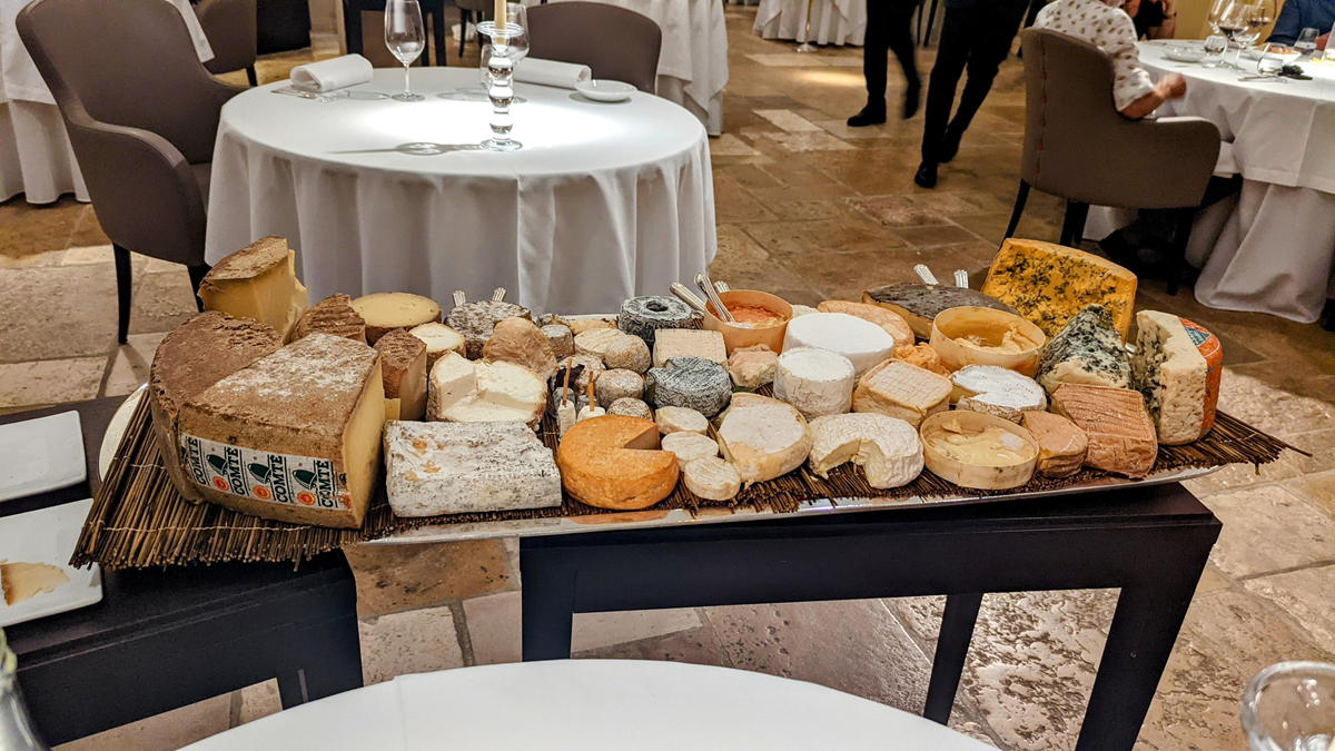 A large assortment of cheeses is displayed on a rectangular platter atop a table in a restaurant setting, with various cheese shapes, colors, and textures surrounded by neatly set dining tables and chairs.