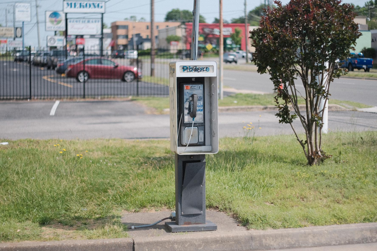 A single payphone stands on a grassy curb beside a street, with cars, shops, and signs visible in the blurred background. Habits.