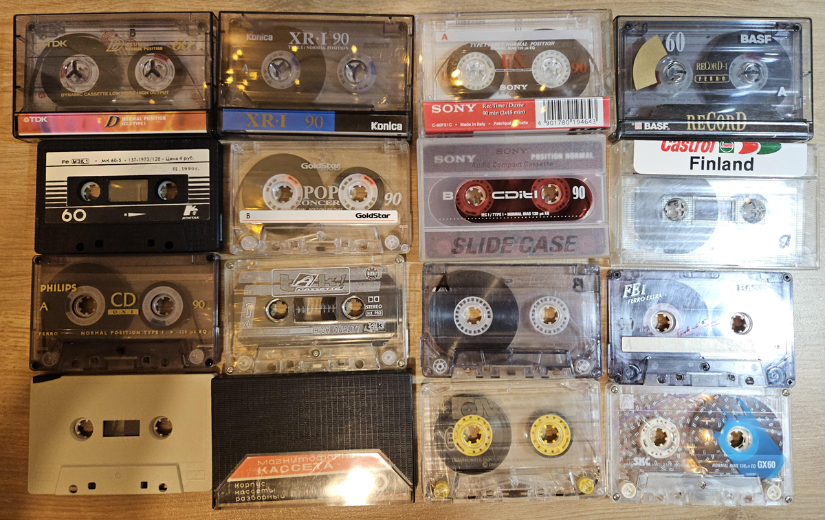 Sixteen assorted cassette tapes, some labeled and some clear, are arranged neatly in a 4x4 grid on a wooden surface. Brands and handwritten notes are visible on several tapes.