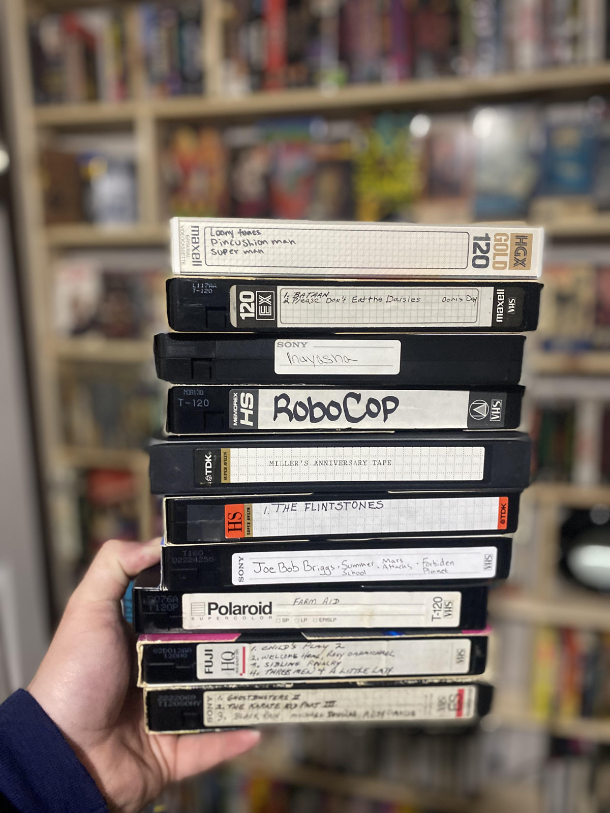 A hand holds a stack of labeled VHS tapes in front of shelves filled with various VHS tapes, with some handwritten and some printed labels visible.
