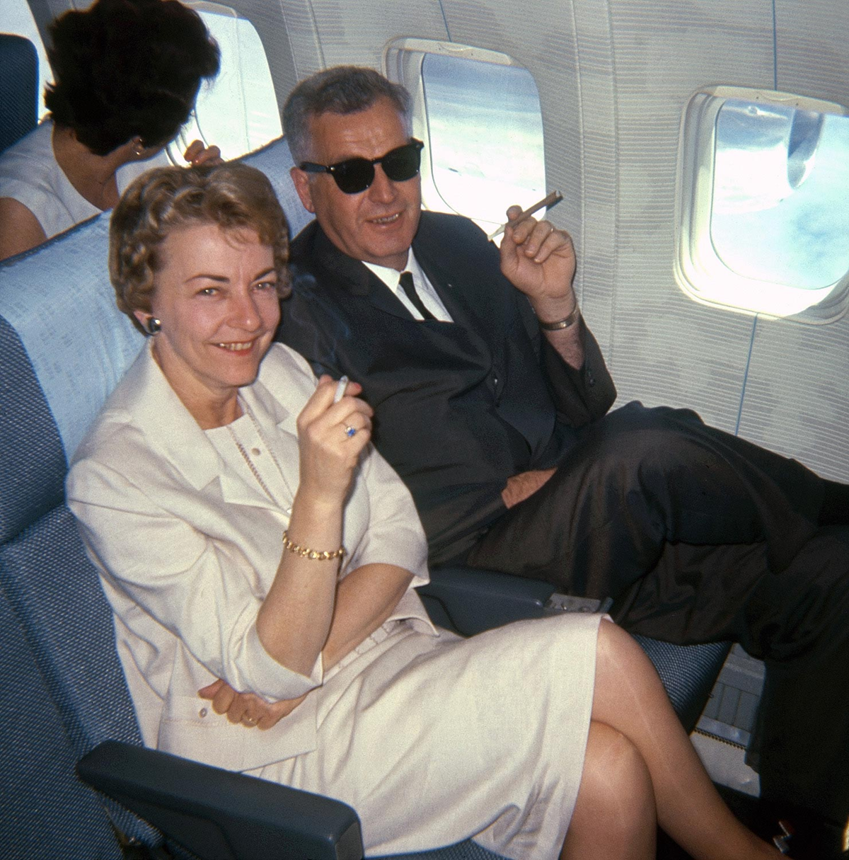 A man and woman sit side by side on an airplane, both smiling and holding cigarettes. The man wears sunglasses and a suit; the woman wears a light-colored dress. Another woman sits in the row ahead, facing away.