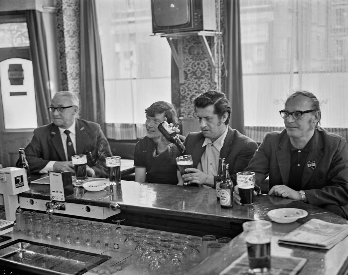Four people sit at a bar counter with drinks in front of them, appearing to converse in a relaxed, well-lit pub setting. Glasses of beer and a bottle are visible on the bar.