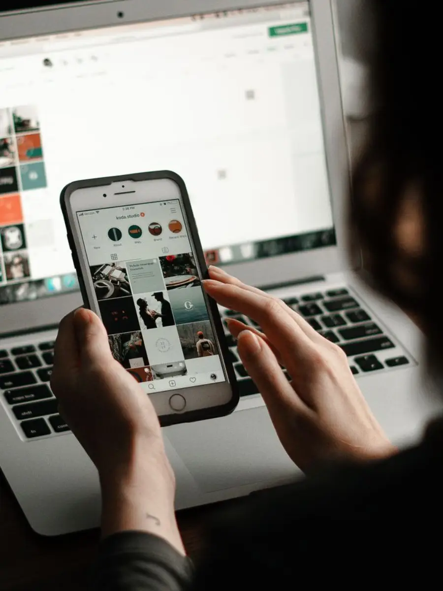 A person uses a smartphone to browse social media, while sitting in front of an open laptop displaying a website.