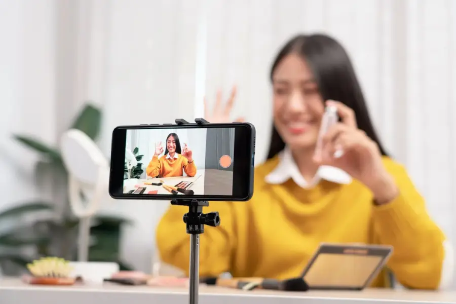 A woman in a yellow sweater sits at a table with makeup products, smiling and waving while recording herself with a smartphone on a tripod.