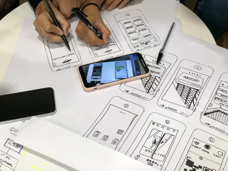 Two people sketch mobile app wireframes on paper with pens. A smartphone lies on top of the sketches, displaying additional wireframe designs. Another phone and more papers are also on the white table.