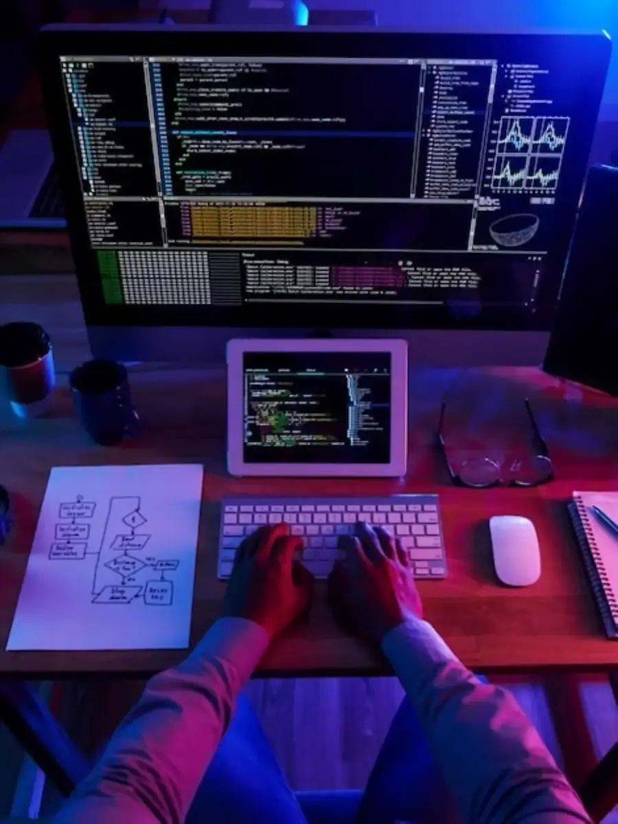 A person types on a keyboard at a desk with a tablet and two monitors displaying code. Nearby are a notepad, mouse, cups, and a paper with a flowchart. The lighting is a mix of blue and purple tones.