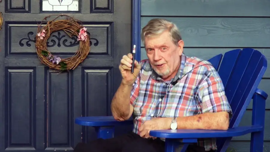An older man with gray hair and a plaid shirt sits in a blue chair on a porch, holding up a vape pen and smiling. A dark blue door with a wreath is visible behind him.