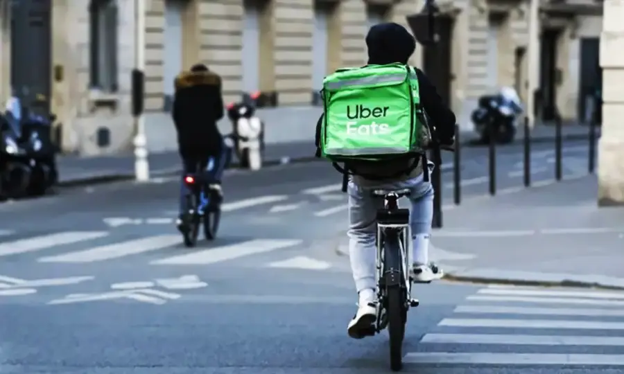 A person wearing a green Uber Eats backpack rides a bicycle on a city street; another cyclist is ahead in the background.