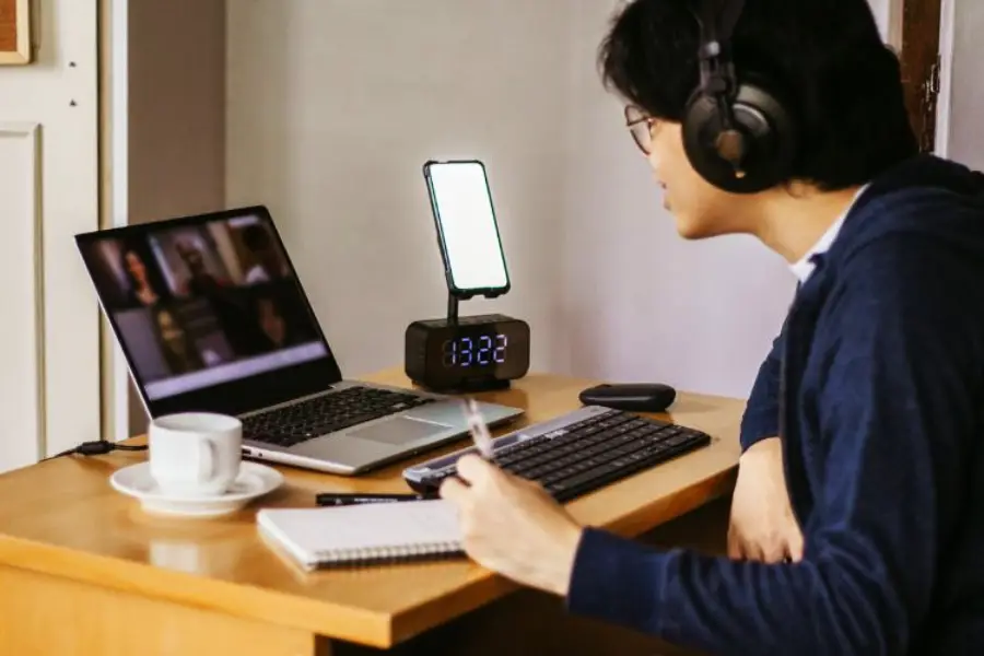 A person wearing headphones sits at a desk with a laptop, smartphone, notebook, pen, and a cup of coffee, appearing to participate in a video call or online meeting.