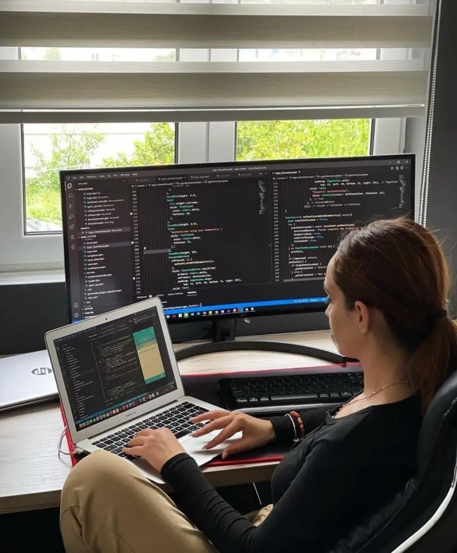 A person is sitting at a desk working on a laptop and a large monitor, both displaying code editors. The scene is set by a window with blinds partially open, letting in natural light.