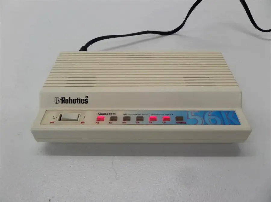A beige USRobotics external modem with indicator lights and a blue "56K" label on the front, connected by a black power cord, sitting on a light gray surface.