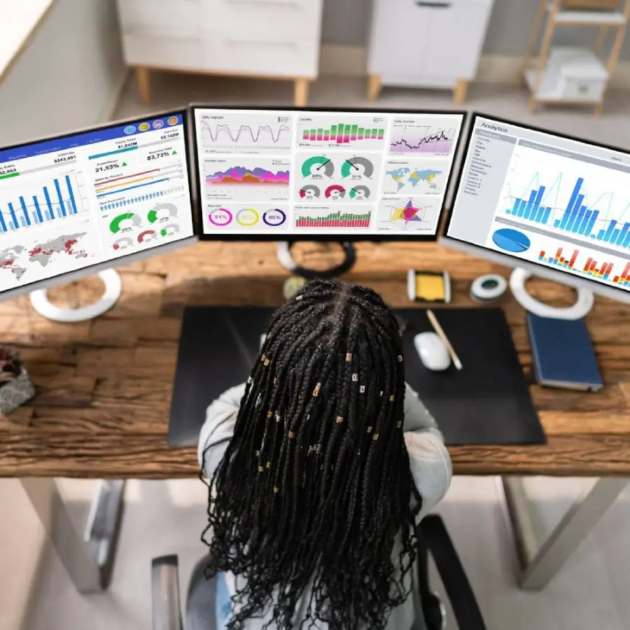 A person with braided hair sits at a desk, facing three computer monitors displaying colorful graphs, charts, and world maps. The workspace includes a keyboard, mouse, phone, and office supplies.