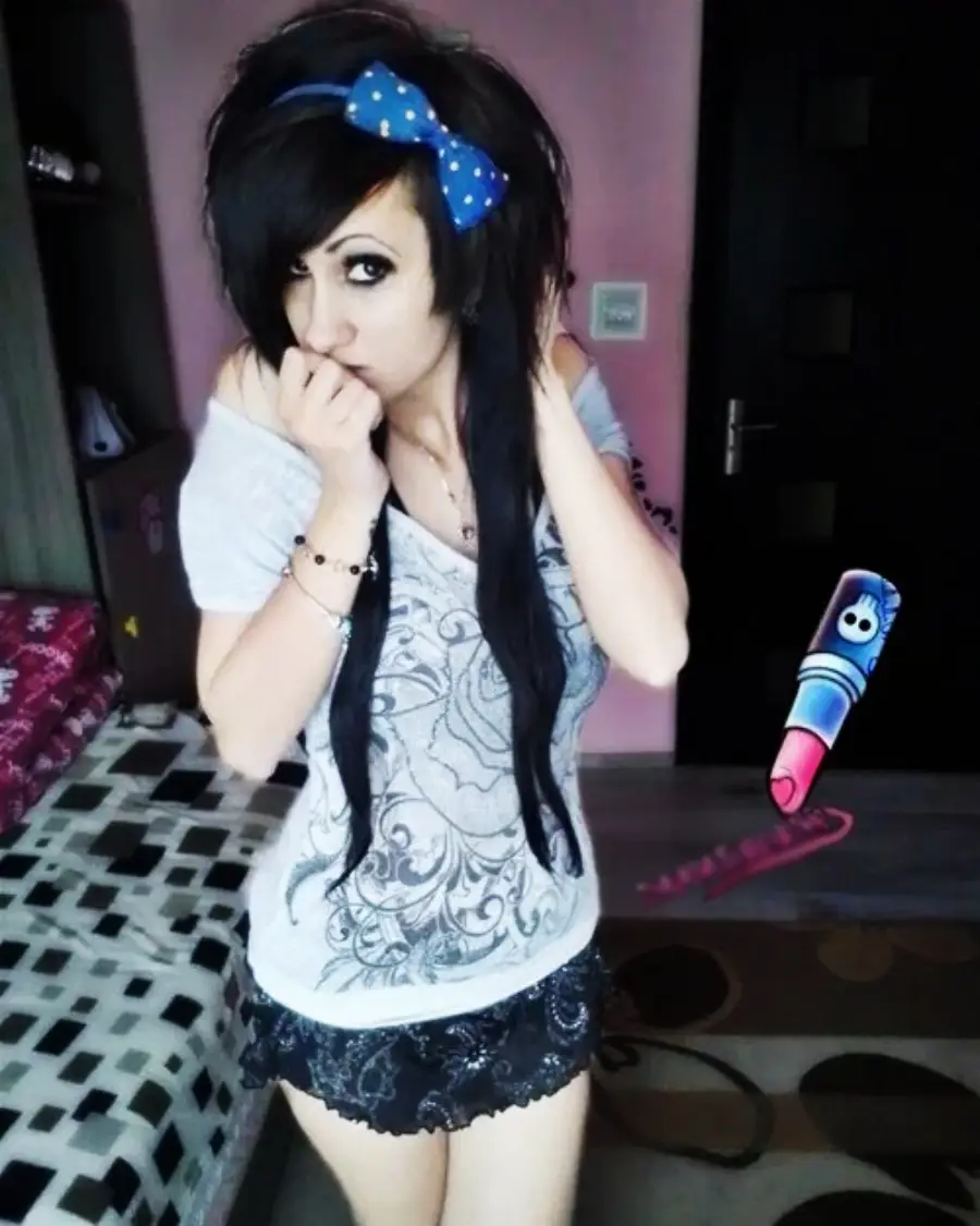 A young woman with long black hair, a blue polka-dot bow, and heavy eyeliner stands in a bedroom, wearing a printed top and a short skirt. She is posing shyly with her hand near her face. A cartoon crayon is edited into the image.