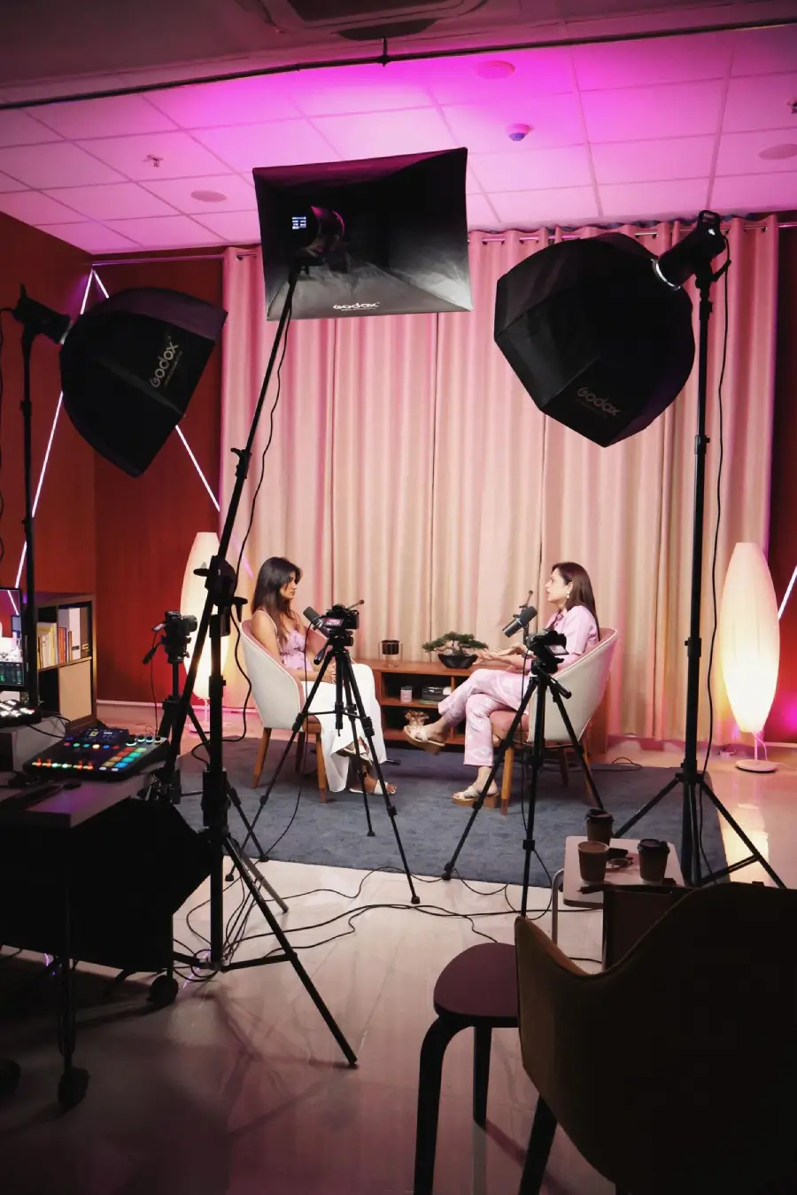 Two women sit across from each other in a studio with microphones, surrounded by lighting equipment and cameras, recording a podcast or interview. The room has pink lighting and modern decor.