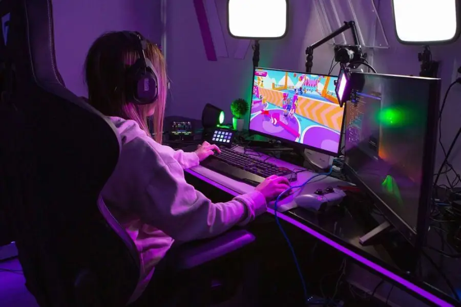 A person wearing headphones sits at a desk playing a colorful video game on a dual-monitor setup, surrounded by gaming equipment and illuminated by purple lighting.