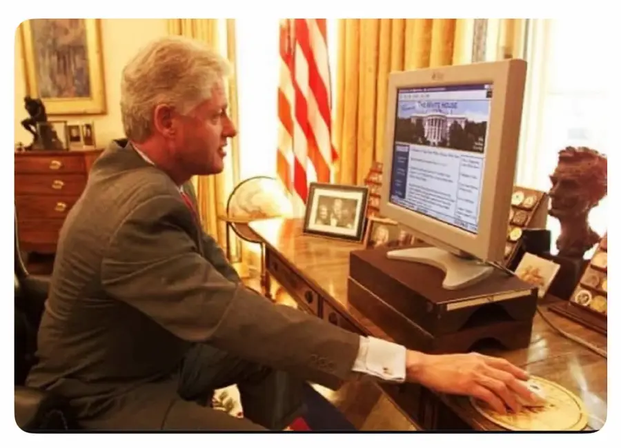 A man in a suit sits at an office desk, using a computer. The monitor displays a website with a White House image. An American flag, framed photos, and a bust are visible in the background.