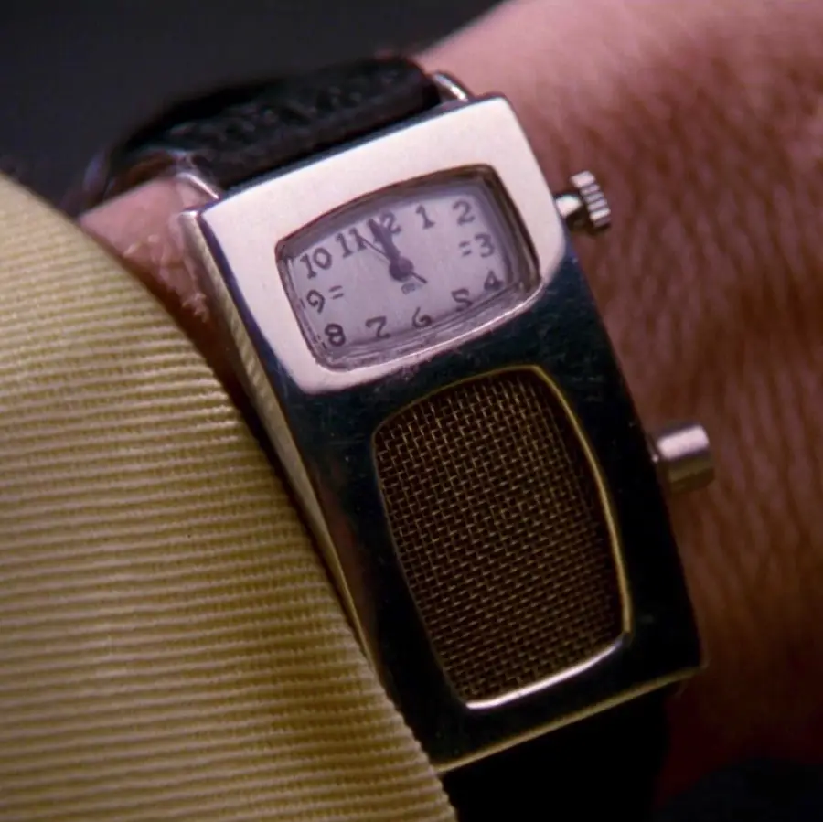 A close-up of a wristwatch with a rectangular face showing 12:00 and a built-in speaker below the dial, worn on a person’s wrist with a yellow shirt sleeve visible.