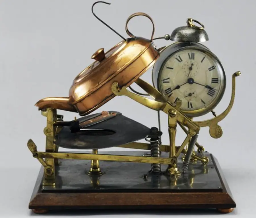 A vintage mechanical alarm clock with a frying pan and spatula attachment designed to cook food, likely eggs, when the alarm rings.