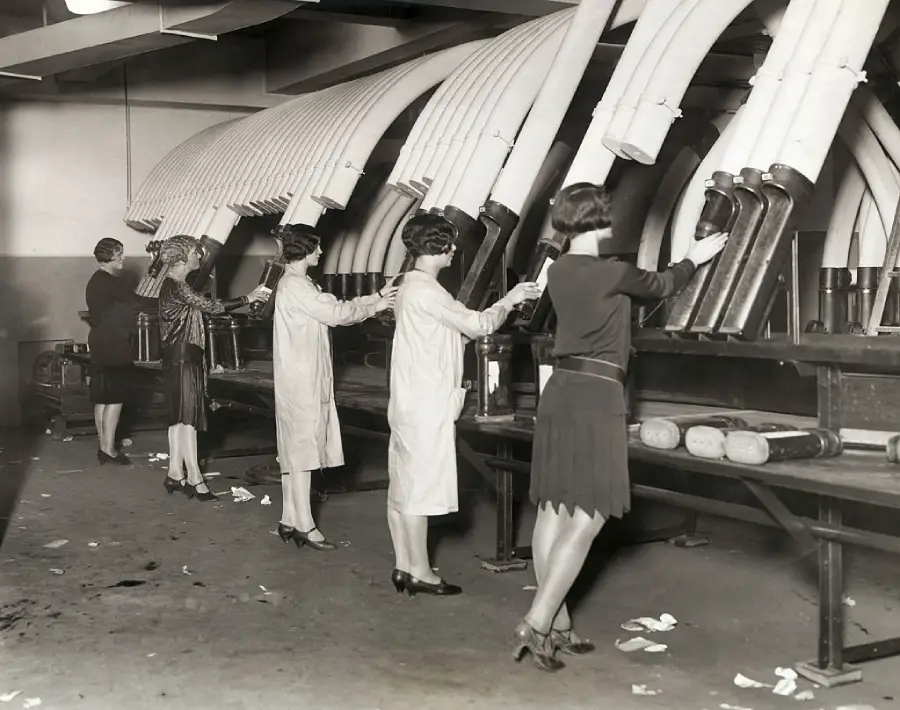Four women work at a conveyor belt in a factory, each handling objects delivered by large tubes overhead. The floor is cluttered with paper, and the women wear dresses and uniforms, suggesting an early-to-mid 20th-century setting.