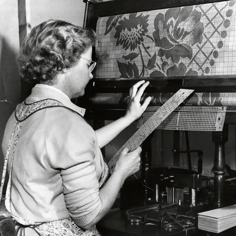 A woman operates a textile loom, holding a punched card while examining patterned fabric being produced by the machine.