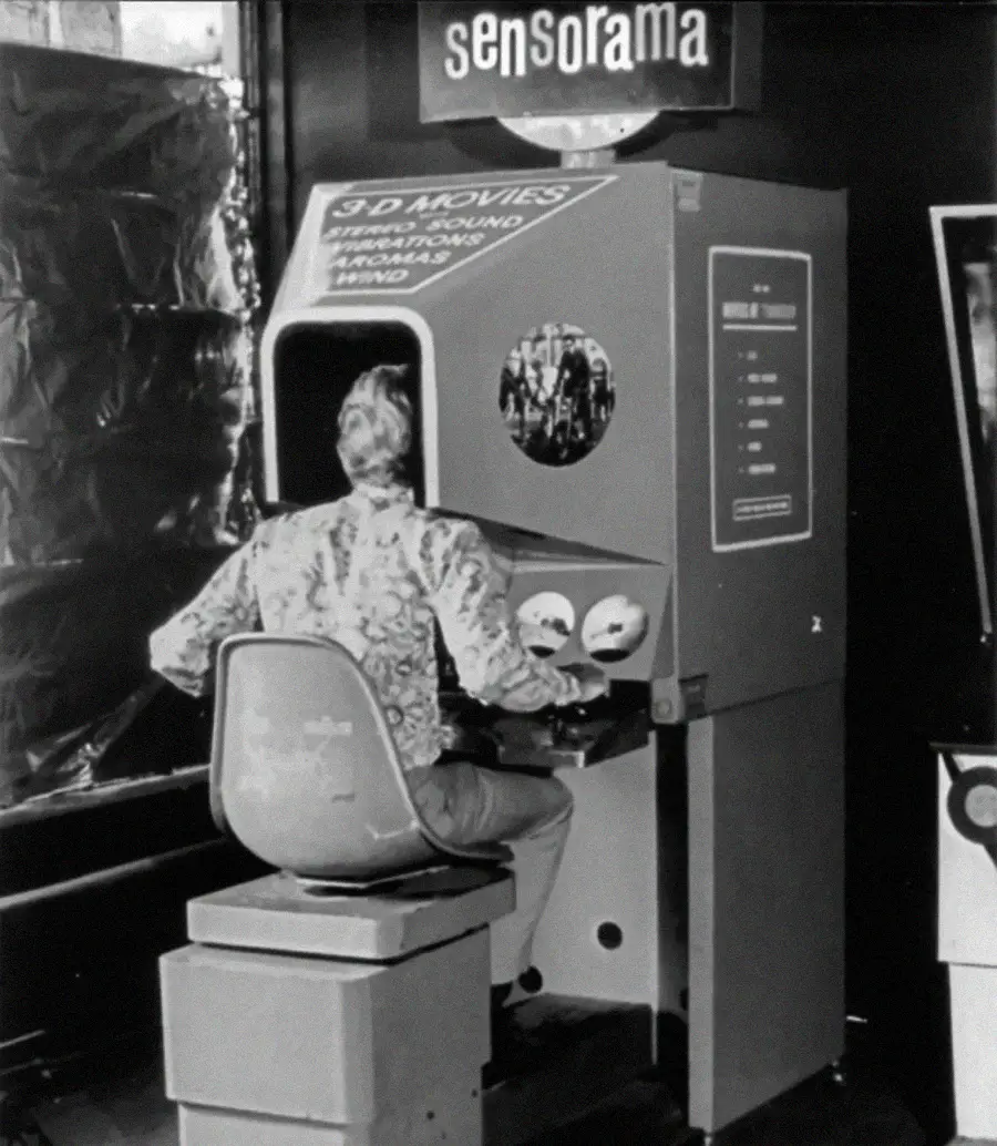 A person sits at the vintage Sensorama machine, wearing headphones and looking into the device, which promises 3D movies with stereo sound, aroma, wind, and vibration effects.