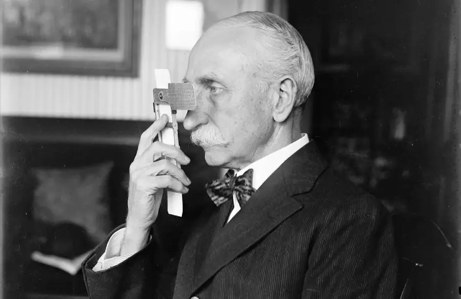 An older man in a suit uses an early vision testing device, holding it up to his eyes. He has a mustache and is seated indoors, viewed in profile.