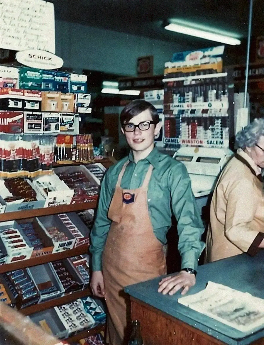 A young person in glasses and a tan apron stands behind a counter in a vintage convenience store, surrounded by shelves of cigarettes and candy. An older woman is at the cash register in the background.