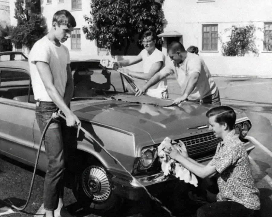 Four young men wash a vintage car outdoors; one uses a hose, another wipes the hood, and two more clean the windshield and side, all appearing focused on the task. Trees and a building are in the background.