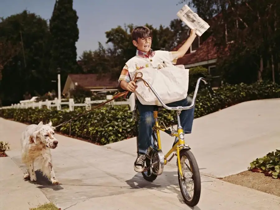 A smiling boy rides a yellow bicycle with one hand while delivering newspapers and holding a paper up. A white dog runs beside him on a leash, and houses with trees are in the background.