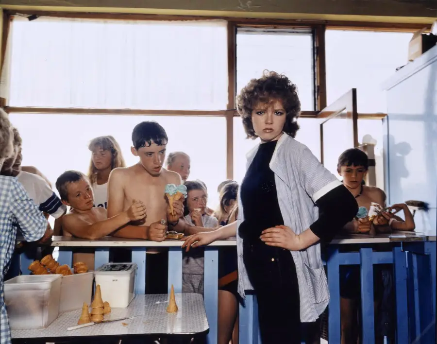 A woman stands confidently at an ice cream counter as several children, some shirtless, hold ice cream cones and wait behind a blue railing. Sunlight streams through large windows in the background.