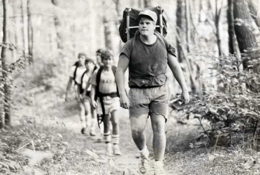 Four boys hike through a wooded trail, each carrying backpacks. The boy in front wears a cap, t-shirt, and shorts, leading the group as they walk among trees and foliage. The photo appears black and white or vintage.