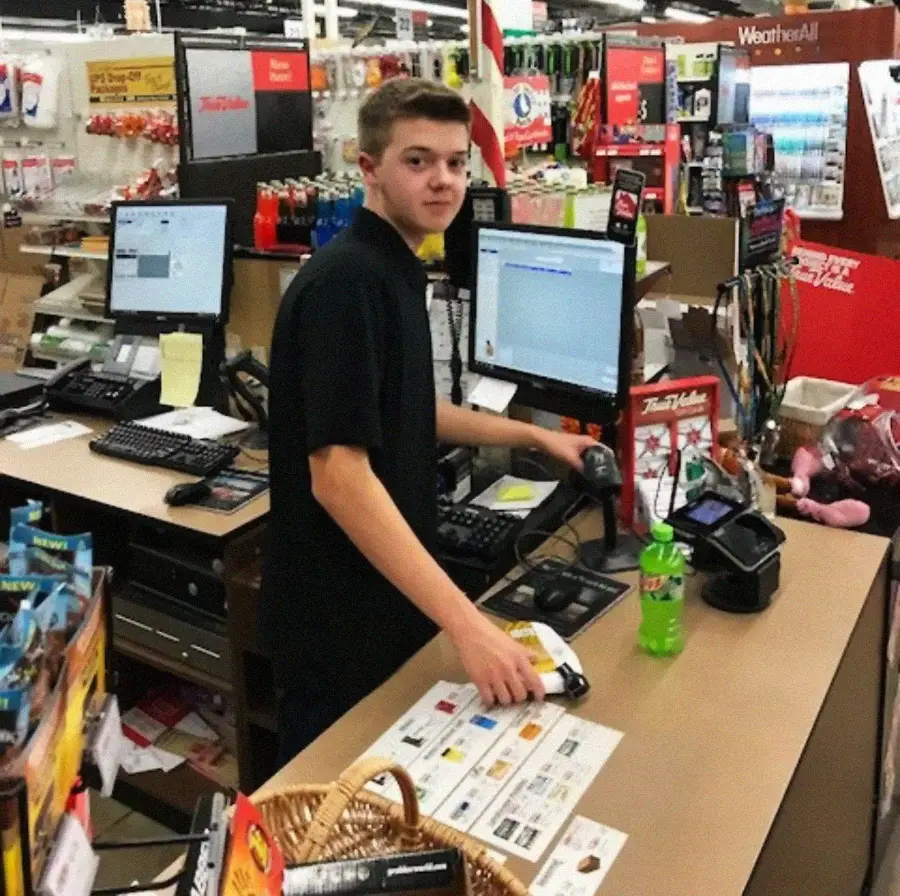 A young cashier in a black shirt stands behind a store checkout counter, using a scanner. There are two computer monitors, a Mountain Dew bottle, candy, and various items on the counter in a busy retail environment.