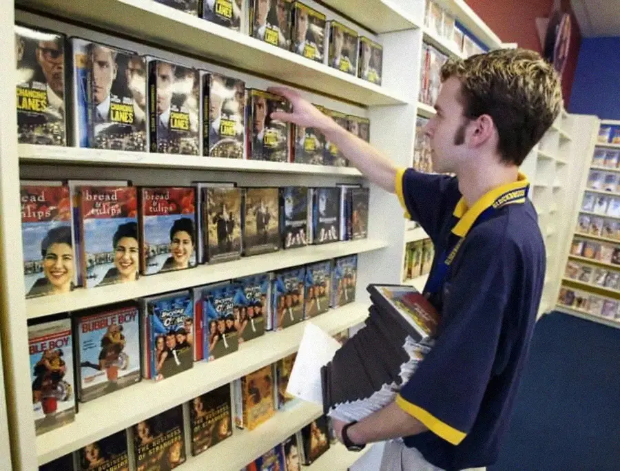 A person in a blue uniform is shelving DVD cases in a video rental store, surrounded by shelves filled with various movie covers.