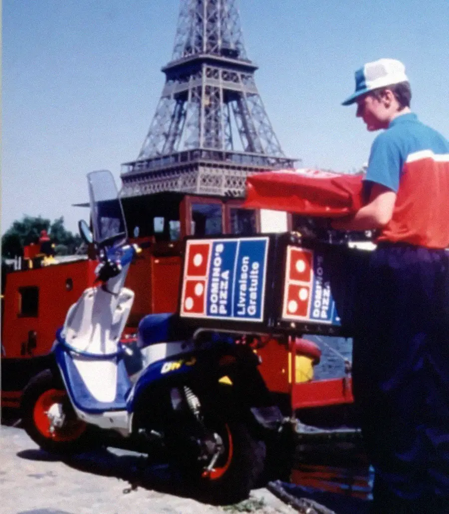 A Domino’s Pizza delivery person stands beside a scooter with delivery boxes, near the Eiffel Tower in Paris, with a red boat in the background.