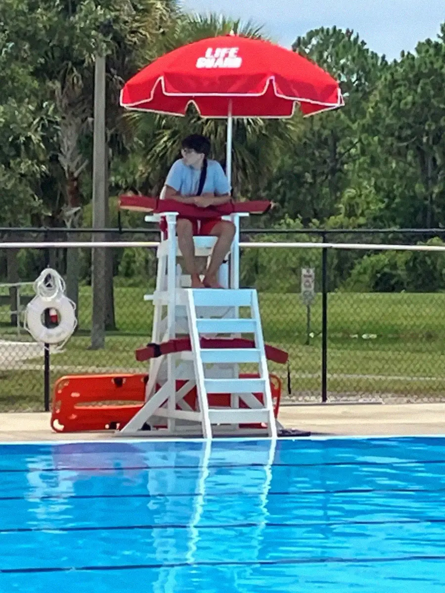 A lifeguard sits on a tall white chair under a red umbrella labeled "LIFEGUARD," watching over an outdoor pool. Trees and a fence are visible in the background.