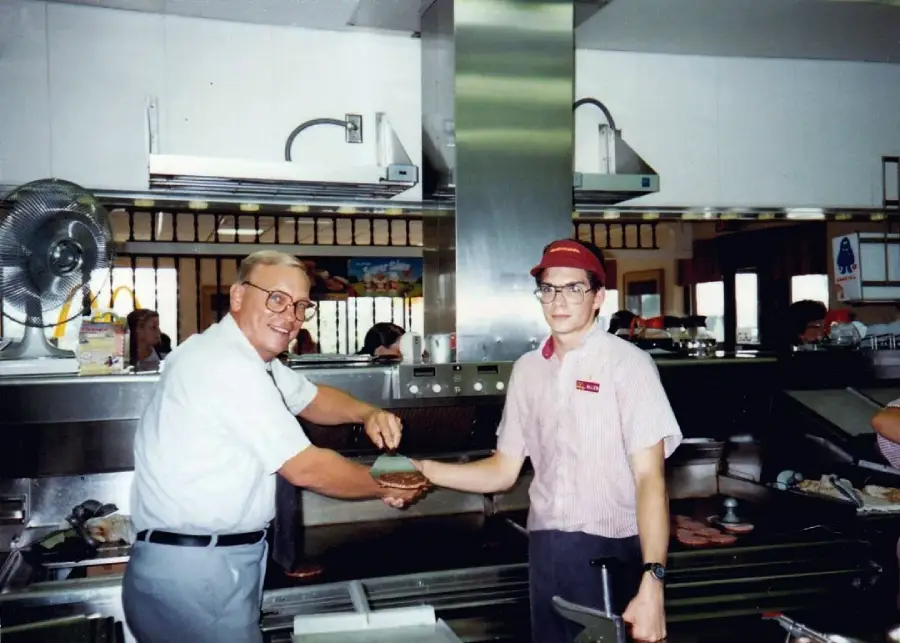 An older man in glasses hands a burger patty to a young fast food worker in uniform behind a restaurant counter, with kitchen equipment and people dining in the background.