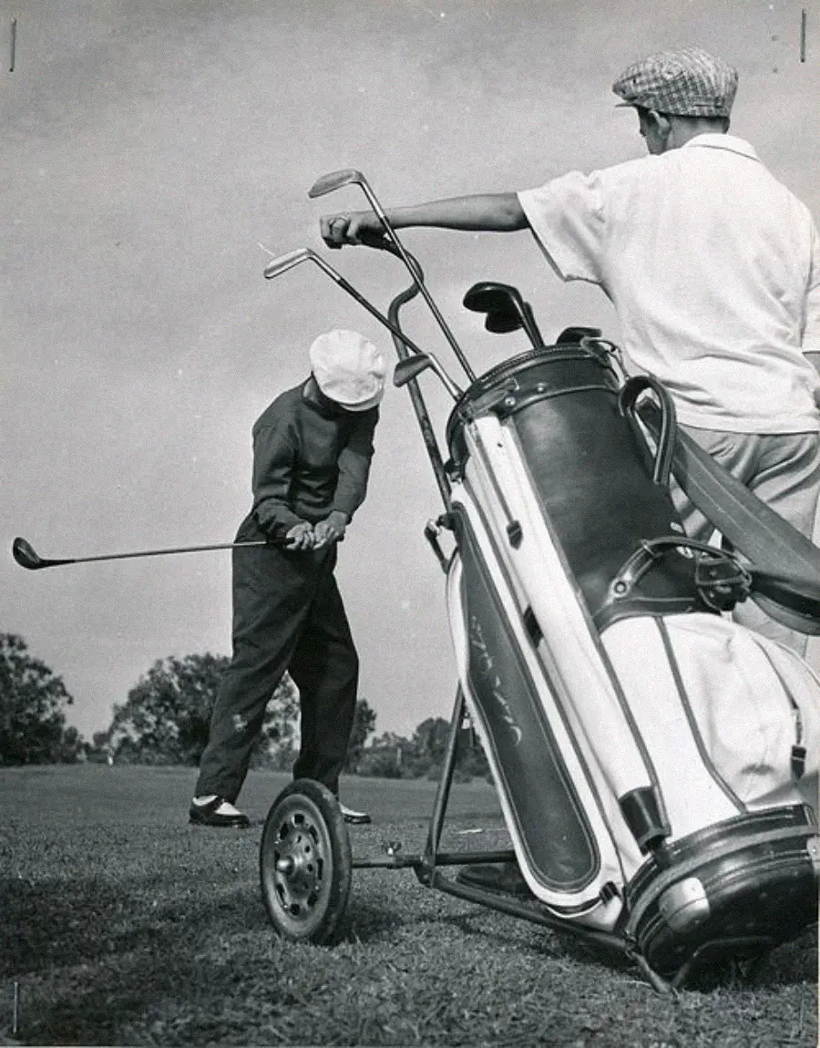 Two golfers on a course: one swings a club while the other stands nearby, resting an arm on a large golf bag attached to a wheeled cart. Both wear hats and vintage clothing. Trees are visible in the background.